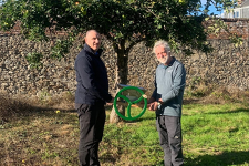 Two men holding a green circular metal device, an apple presser, standing in front of an apple tree