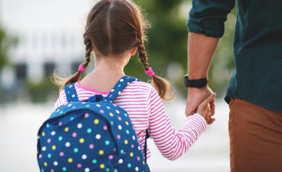 Photograph from behind of a young girl wearing a backpack and holding her father/male carer's hand